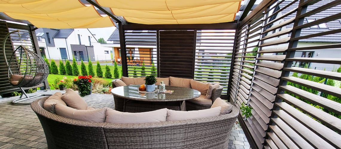 Eleganckie drewniane zadaszenie tarasu z kompletem mebli ogrodowych, w tym okrągłym stołem i wygodnymi sofami, w nowoczesnym ogrodzie przydomowym.