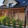 Drewniana pergola z zadaszeniem na tarasie przy domu jednorodzinnym otoczona ozdobnymi trawami.