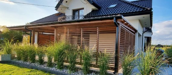 Drewniana pergola z zadaszeniem na tarasie przy domu jednorodzinnym otoczona ozdobnymi trawami.