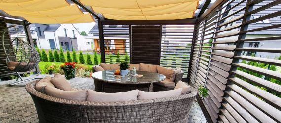 Eleganckie drewniane zadaszenie tarasu z kompletem mebli ogrodowych, w tym okrągłym stołem i wygodnymi sofami, w nowoczesnym ogrodzie przydomowym.